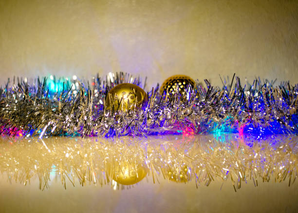 Christmas balls, hidden in Christmas tinsel, with colorful lights from the garland in tinsel on a glass surface with reflection. New Year's composition