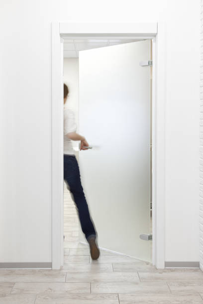Person entering a room in office with minimalist white interior thru frosted glass door Young woman entering a room in modern office with minimalist white interior through frosted glass door. Woman is motion blurred frosted glass door office stock pictures, royalty-free photos & images