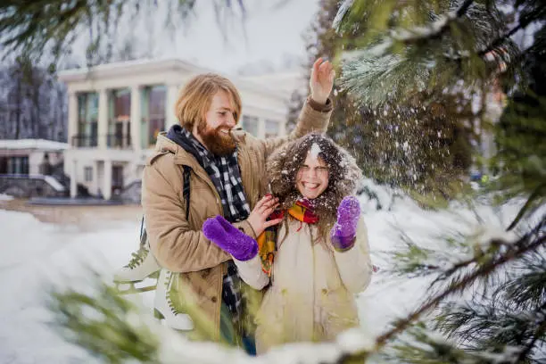 Young loving couple Caucasian man with blond long hair and beard, woman with toothy smile have fun, indulge, fool around winter park near Christmas tree, coniferous tree after ice skating in winter.