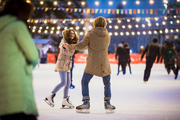 giovane coppia innamorata dell'uomo caucasico con i capelli biondi con i capelli lunghi e la barba e bella donna si divertono, pattinaggio attivo sulla scena del ghiaccio nella piazza della città in inverno la vigilia di natale - ice skates foto e immagini stock