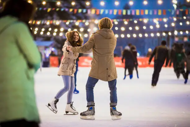 Young couple in love Caucasian man with blond hair with long hair and beard and beautiful woman have fun, active date ice skating on the ice arena in the evening city square in winter on Christmas Eve