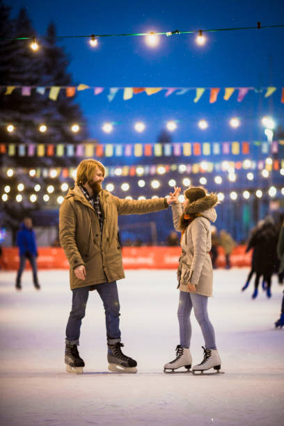 jeune couple amoureux du caucase l’homme aux cheveux blonds aux cheveux longs et barbe et belle femme ont date de fun, active, patiner sur la scène de la glace à la place de la ville en hiver le soir de noël - pair couple love humor photos et images de collection