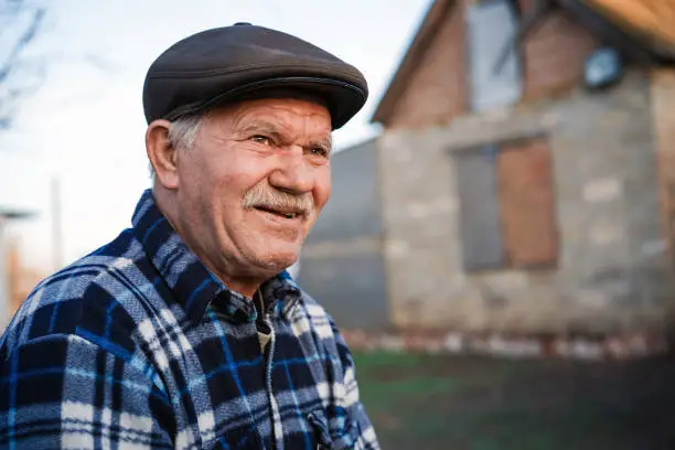 Photo of Happy smiling elder senior man portrait with a mustache in a cap on the background of a brick house in a Russian village