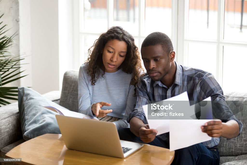 Serious African American couple discussing paper documents Serious African American couple discussing paper documents, sitting together on couch at home, man and woman checking bills, bank account balance, terms of contract, mortgage, loan agreement Couple - Relationship Stock Photo