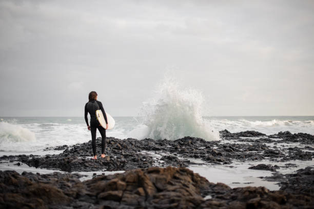 Rear view brunette boy standing with a surf in his hands on the rock beach looking on the wave.