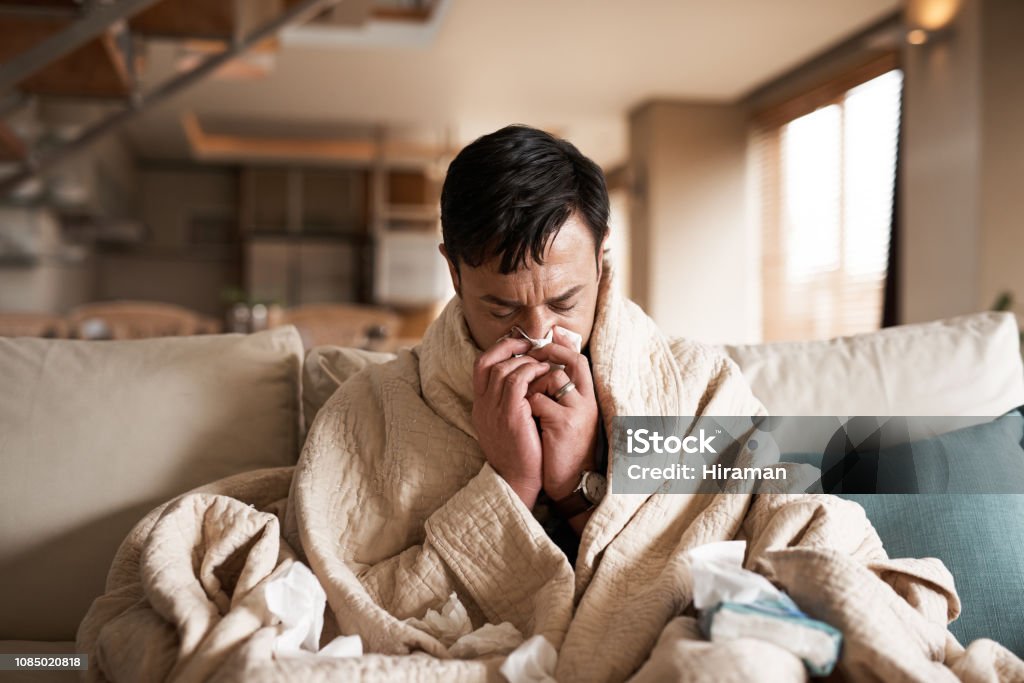 It's the season of sneezes Cropped shot of a young man suffering with flu while sitting wrapped in a blanket on the sofa at home Cold And Flu Stock Photo