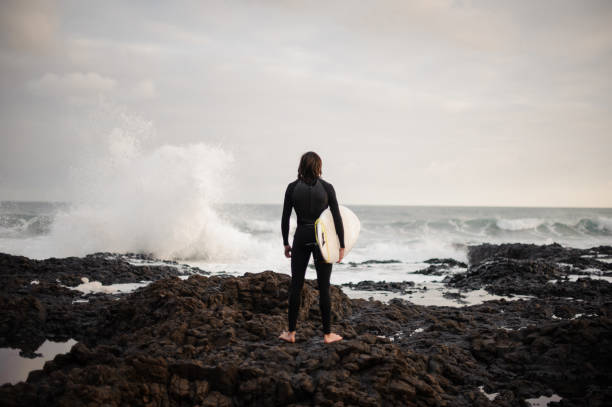 Back view man standing with a white surf in his hands on the pebble beach in the background of the ocean.