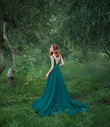 beautiful woman in the forest is standing with her back to the camera. deep cut on the emerald wonderful dress with a long train. red hair in the lights of the sun. looking up to the sky. open back.
