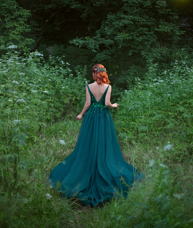 red hair girl is going to the forest. standing with her back to the camera. she is looking for her way. flower in hair. deep cut on the emerald wonderful dress with a long train. no face on the photo.