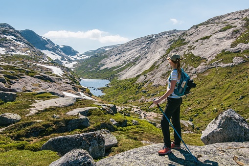 Backpacker traveller girl hikes a trekking trail and stares on beautiful scenic panorama landscape of snowy mountains and lake in Kjerag, Norway