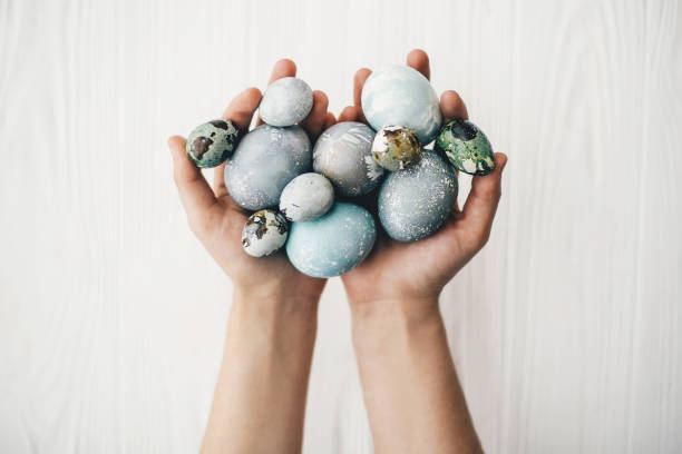 istock Stylish easter eggs in female hands on white wooden background. Hands holding modern easter eggs painted with natural dye in grey and blue marble color. Happy Easter 1085054254