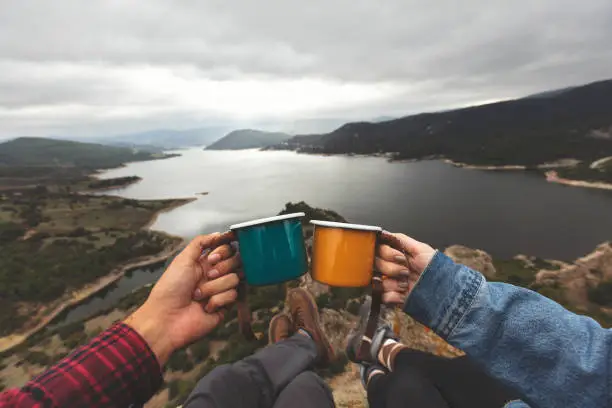Pov image of couple holding enamel cups on mountain peak