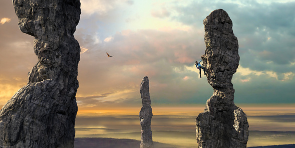 A young woman wearing leggings, top, climbing shoes with chalk bag hanging from a rock face with one arm and foothold, reaches down to her chalk bag. The rock climber is free climbing a tall eroded column rock formation, climbing high up, close to other rock columns under a beautiful dawn sky.