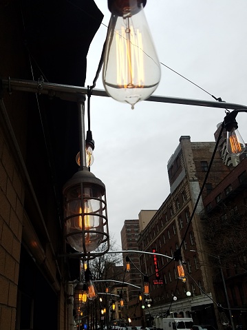 Sidewalk scene. Series of filament pendant lamps hung on wire above. Streetscape at dusk. Relaxed vibe. Sidewalk cafes.
