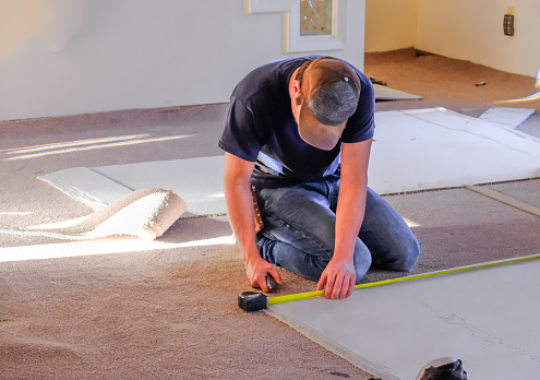 Young man carpenter measuring new rugs he's going to install in house; sunlight coming from the windows