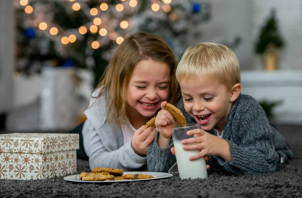 A cute little caucasian brother and sister laugh as they eat cookies and milk together over the holidays.