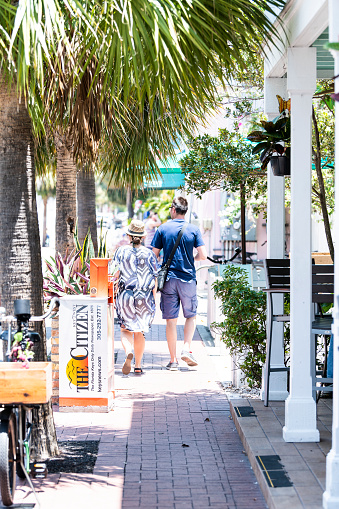 Key West, USA - May 1, 2018: People couple walking on sidewalk of street near restaurant, cafe with the citizen journal news stand, palm trees in Florida city, sunny day on street, parked bicycles