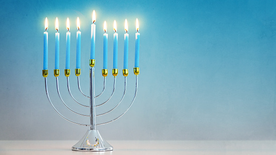 Celebrating Hanukkah with illuminated candles in silver and gold menorah on table with room for text in blue background