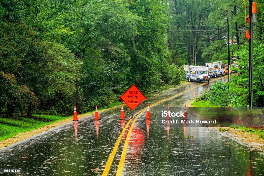 Storm Damage Road Closure A view of a wet curved road closed during a storm with an orange sign and cones while utility trucks wait up the road. Hurricane - Storm Stock Photo