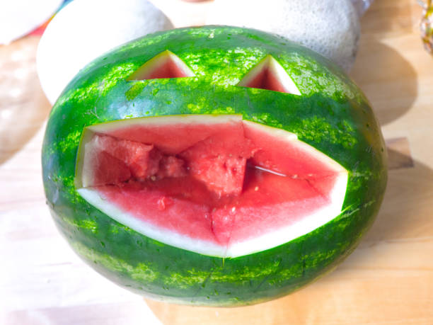 istock Fun carved green oval shaped watermelon face with angular mouth and triangle eyes exposing the pink or red fruit within sitting on a table or counter. 1085133010