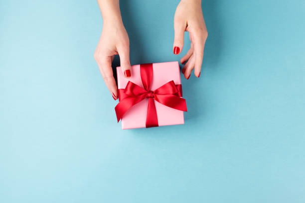 Female hands holding gift box on blue background. Overhead view on female hands with pink gift box wrapped with red bow on blue background. Minimal styled composition. hand holding gift stock pictures, royalty-free photos & images