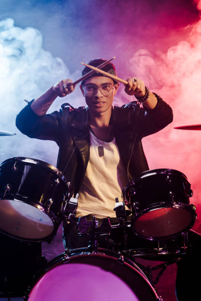 istock happy mixed race male musician raising drum sticks while sitting behind drum set on stage with dramatic lighting and smoke 1085273854