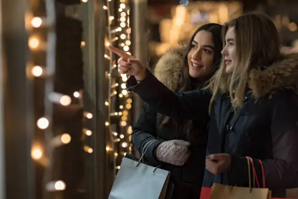 Two smiling women are standing outside a store looking into a window during Christmas time.
