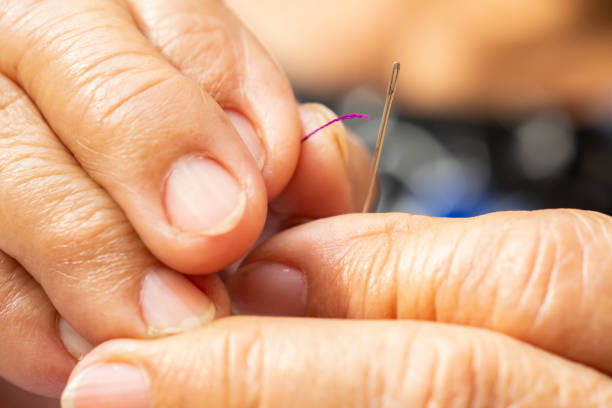 Senior woman's hands trying to thread a needle, Purple colour roll of thread, Close up & Macro shot, Selective focus, Blurred and bokeh background, Tailor, Needlework concept