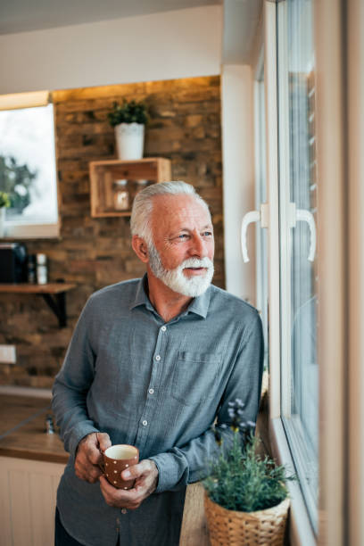 istock Smiling senior man looking through window and drinking coffee at home. 1085333380