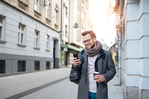 Smiling young man with takeaway coffee and smart phone walking in the town street on winter day.
