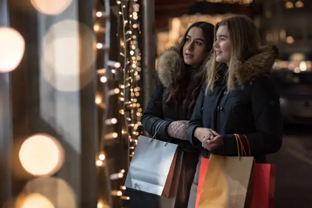 Two smiling women are standing outside a store looking into a window during Christmas time.