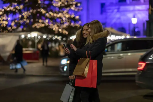 Two young women are looking down at a smartphone. They are holding shopping bags.