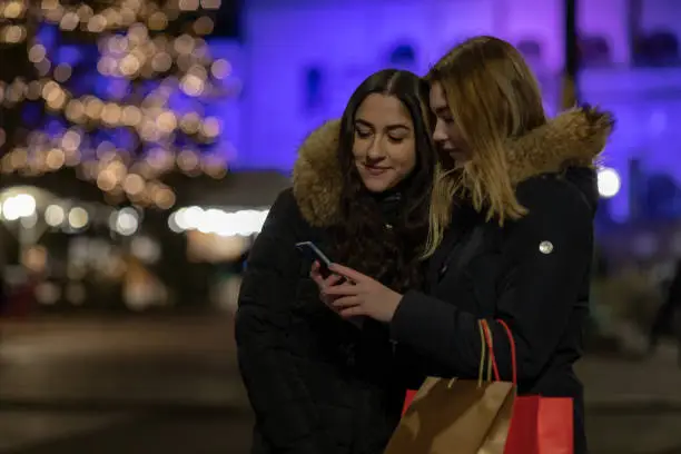 Two young women are looking down at a smartphone. They are holding shopping bags.
