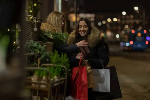 Two smiling women are standing on a street during Christmas time. They are looking at the gifts they have bought.
