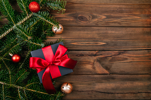 Christmas gift box wrapped in kraft black paper with red ribbon decoration on rustic brown wooden background with fir tree, tree decoration and gold bells. Flatlay style.