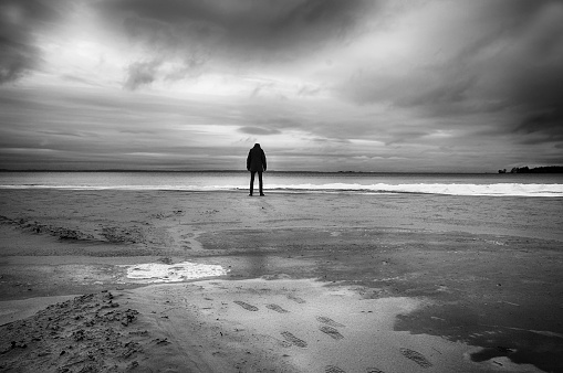 Silhouette of person standing head down on deserted beach in wintertime.
