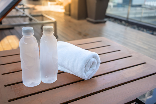 Two drinking bottle of water with white towel on wooden table near swimming pool.