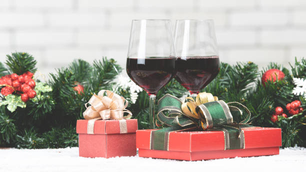 istock Two red wine glasses set next to red gift boxex on snowy table prepare for celabraion, decorate with pine branches and holly berries. Blurred white brick wll copy space at top part 1085504184