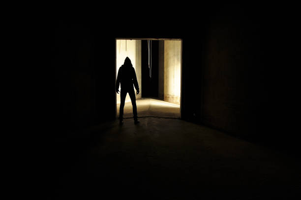 suspicious looking silhouette of hooded person in doorway. unrecognizable silhouette of person with a hood standing in a door way of an empty concrete room. haunted house stock pictures, royalty-free photos & images