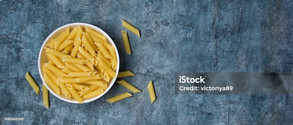 Top view on italian raw penne rigate pasta in wooden bowl Top view of wide textured blue concrete banner background with italian raw penne rigate pasta in wooden bowl with copy space Backgrounds Stock Photo