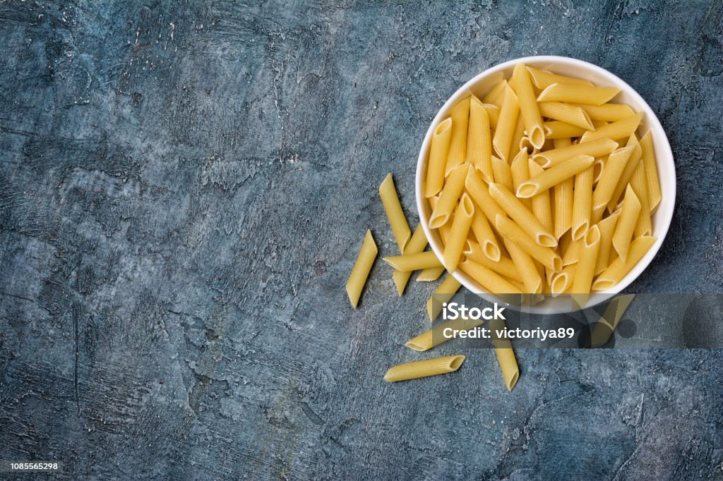 Top view on italian raw penne rigate pasta in wooden bowl Top view on italian raw penne rigate pasta in wooden bowl on blue concrete background with copy space Backgrounds Stock Photo