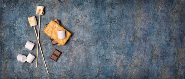 Top view on sticks with roasted on campfire marshmallow, crackers and chocolate as ingredients for s'mores Top view on sticks with roasted on campfire marshmallow, crackers and chocolate as ingredients for s'mores on blue concrete background with copy space smore photos stock pictures, royalty-free photos & images