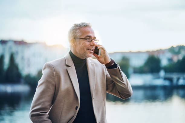 istock Mature businessman with smartphone standing by river at sunset, making phone call. 1085567720