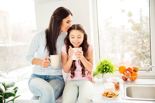 Charming adorable attractive beautiful caucasian kind young smiling mum kissing her small little offspring daughter, wearing casual, sitting on countertop table, drinking cacao in light kitchen