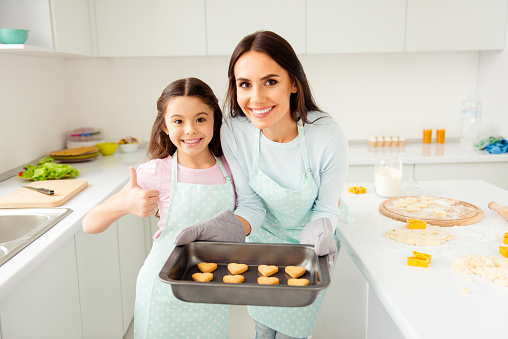 Charming adorable attractive beautiful brunette caucasian kind smiling young mum and her small little offspring daughter, wearing apron, showing thumb up, holding prepared cookies in light kitchen