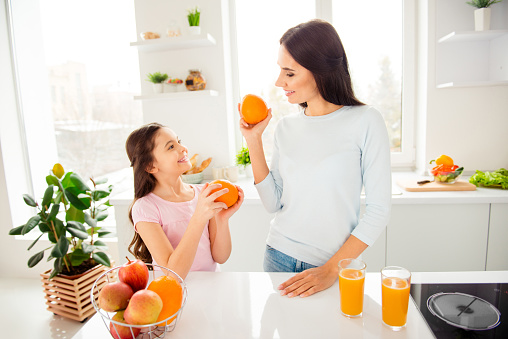 Beautiful lovely nice charming cheerful mom and her small little daughter in casual, holding, showing each other grapefruits, healthy natural orange fresh juice on table in light kitchen