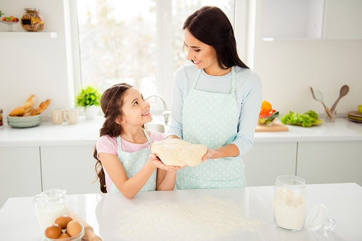 Charming adorable attractive beautiful brunette caucasian kind smiling young mum and her small little schoolgirl daughter wearing apron, preparing cake in light kitchen