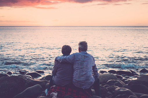 love and romance with adult  matures - elderly couple sitting and hugging each other looking at the sea at sunset relaxing. Concept of vacation, leisure time, relaxation -