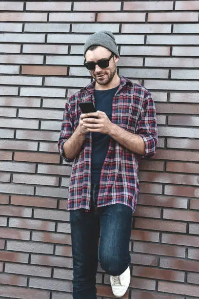 Photo of Typing text message. Side view of handsome young man in smart casual wear holding mobile phone while leaning at the brick wall.