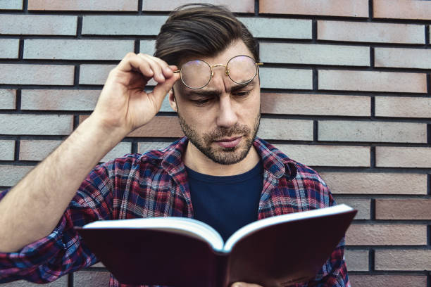 istock Young hipster guy wearing eyeglasses reading a book or note book on brick wall background. 1085616286